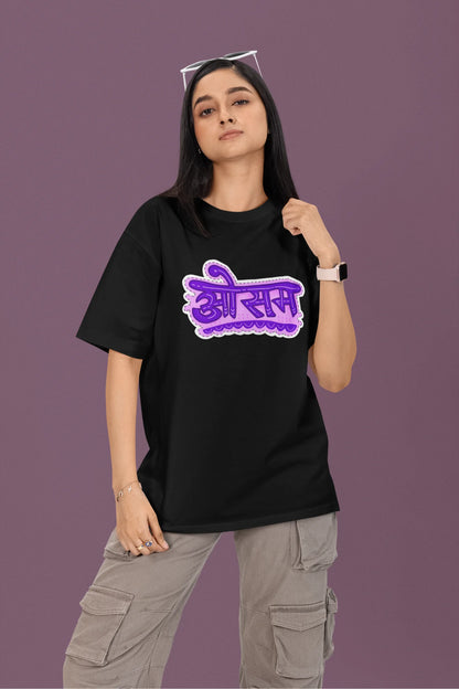 First front view of a female model wearing a black colored oversized graphic t-shirt featuring the word "awesome" in a Hindi script.