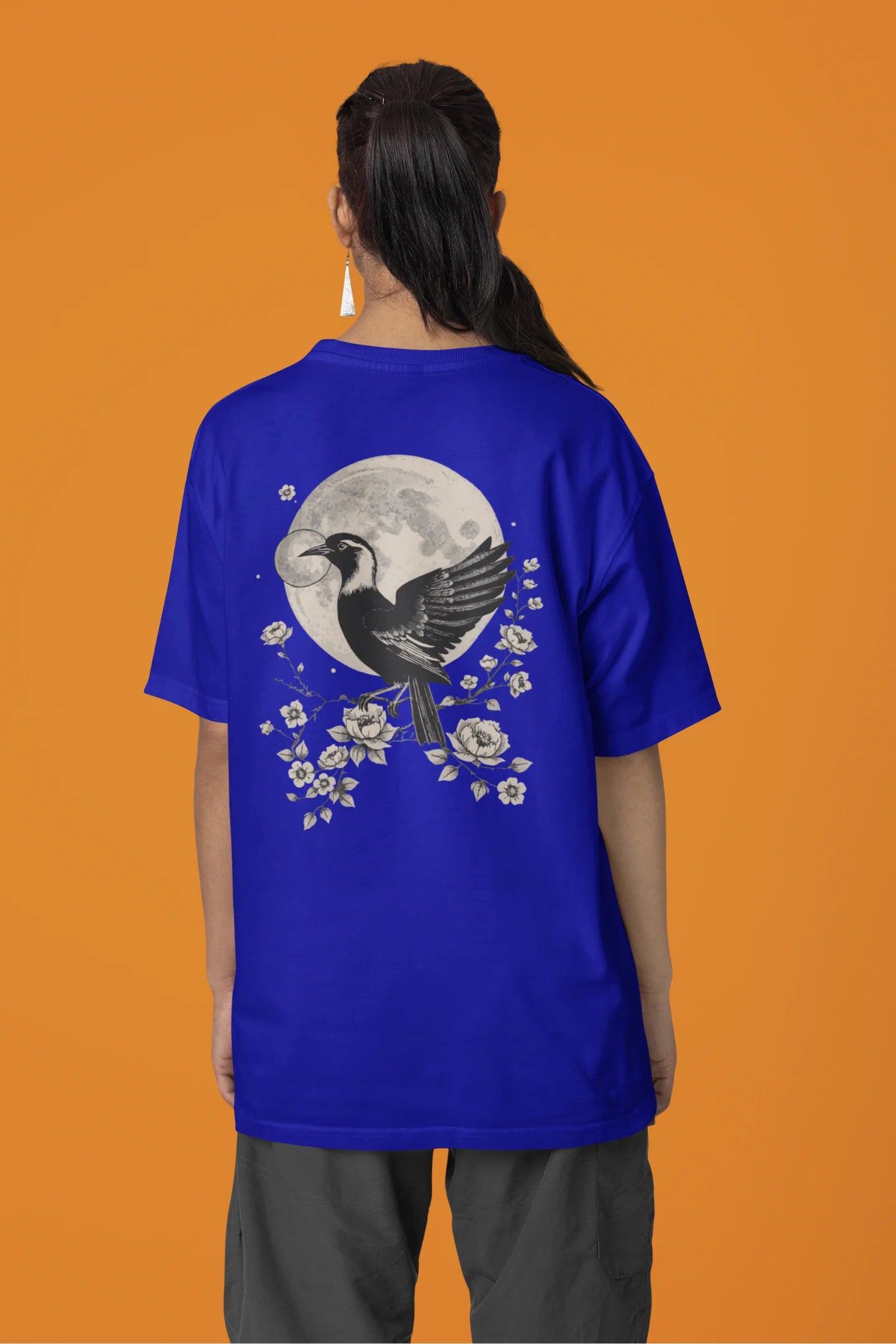 Back view of a female model wearing a royal blue oversized tee with a heron taking off under the moonlight.