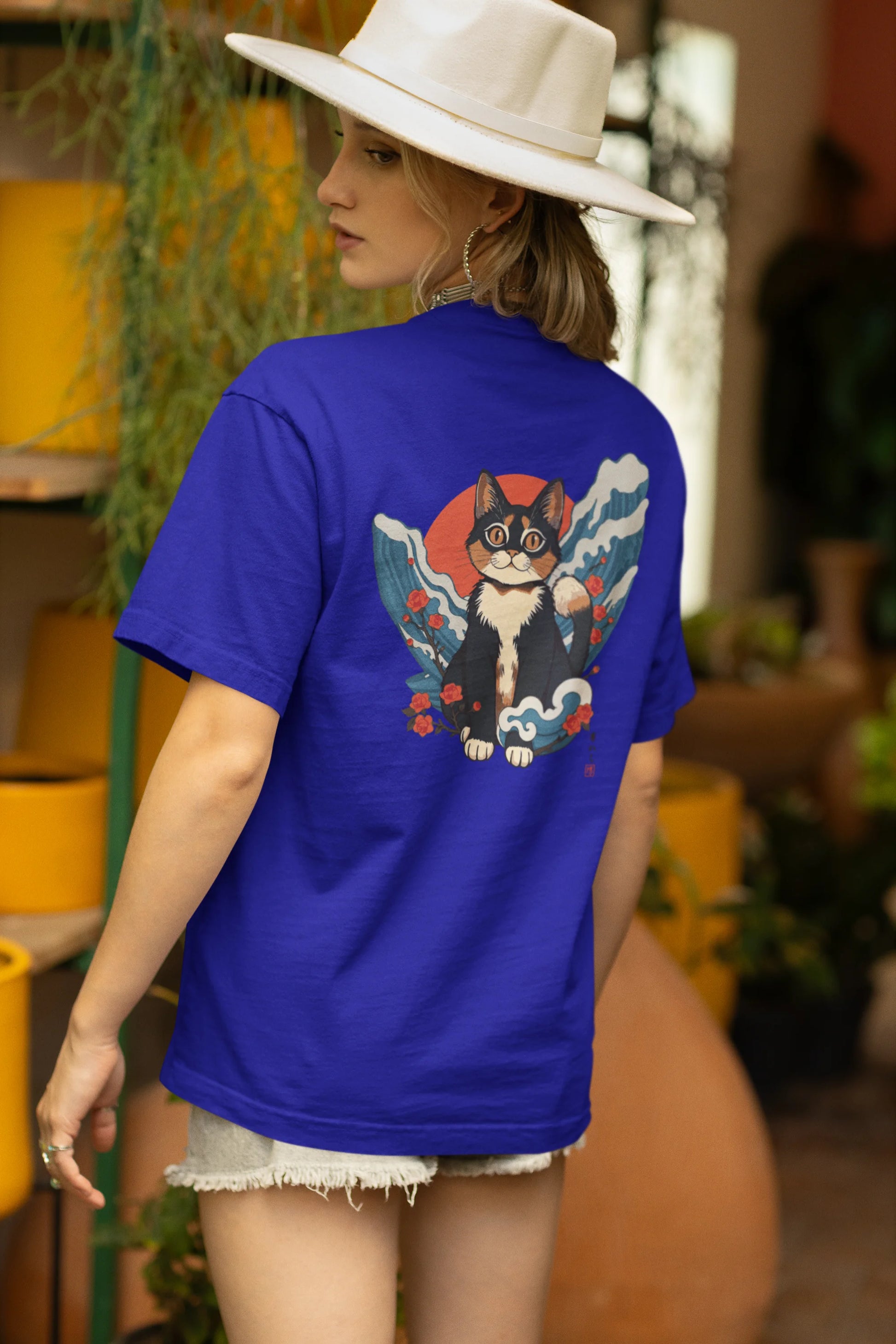 Back view of a female model wearing a royal blue oversized tee with a design of a cat dressed as a samurai, standing against a backdrop of waves and a rising sun.