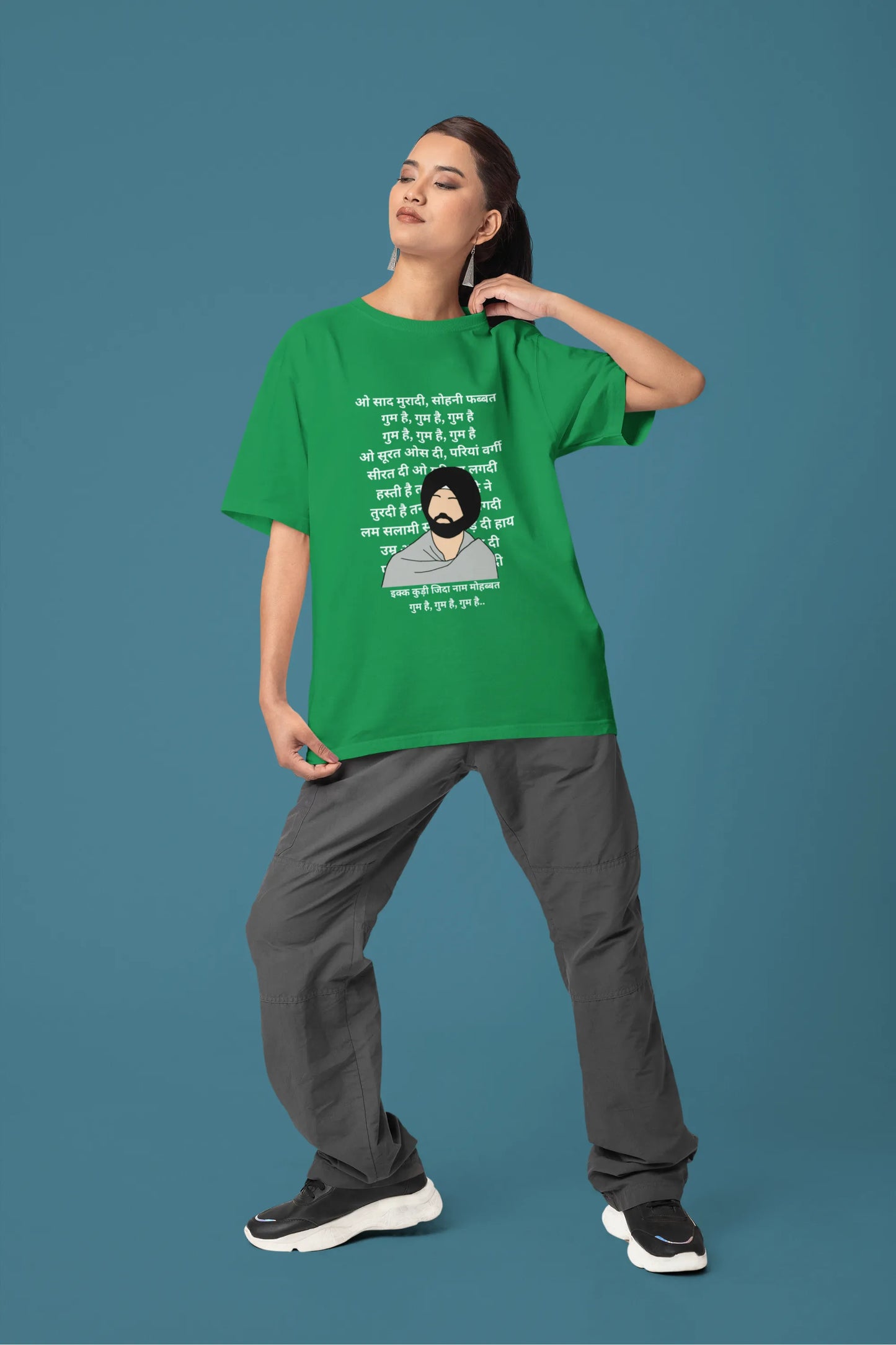 Fifth front view of a female model wearing a forest green oversized  T-shirt with a Diljit Dosanjh portrait  with lyrics from the song "Ikk Kudi" in the background.