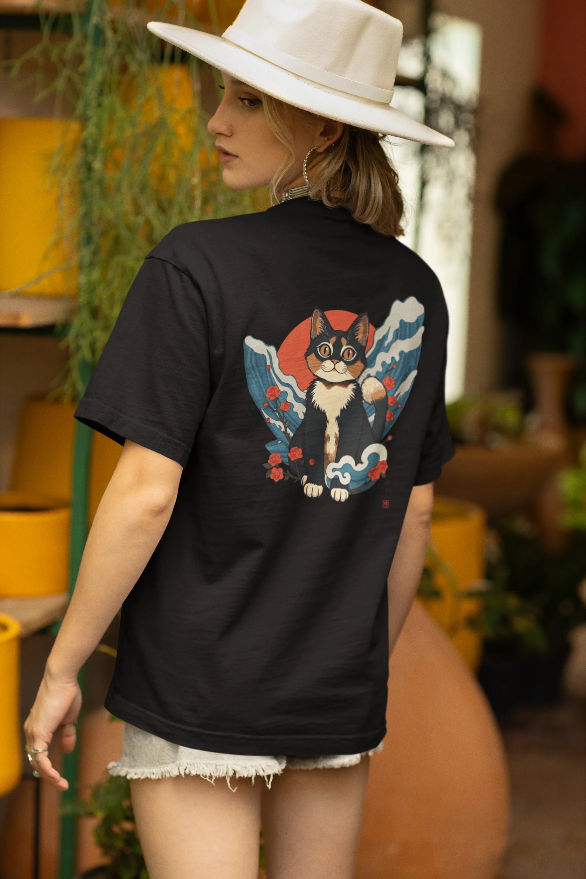 Back view of a female model wearing a black oversized tee with a design of a cat dressed as a samurai, standing against a backdrop of waves and a rising sun.