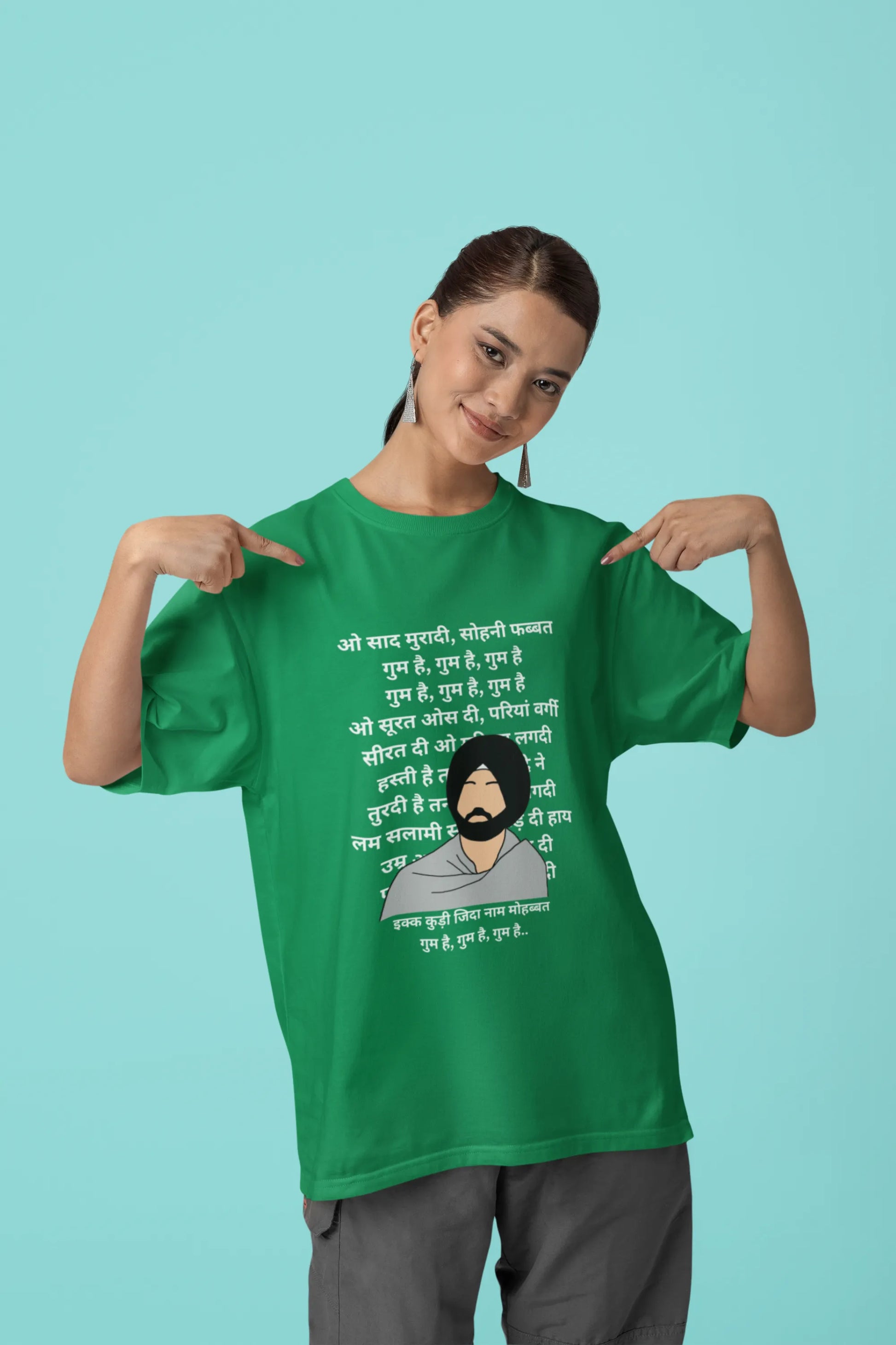 First front view of a female model wearing a forest green oversized  T-shirt with a Diljit Dosanjh portrait  with lyrics from the song "Ikk Kudi" in the background.