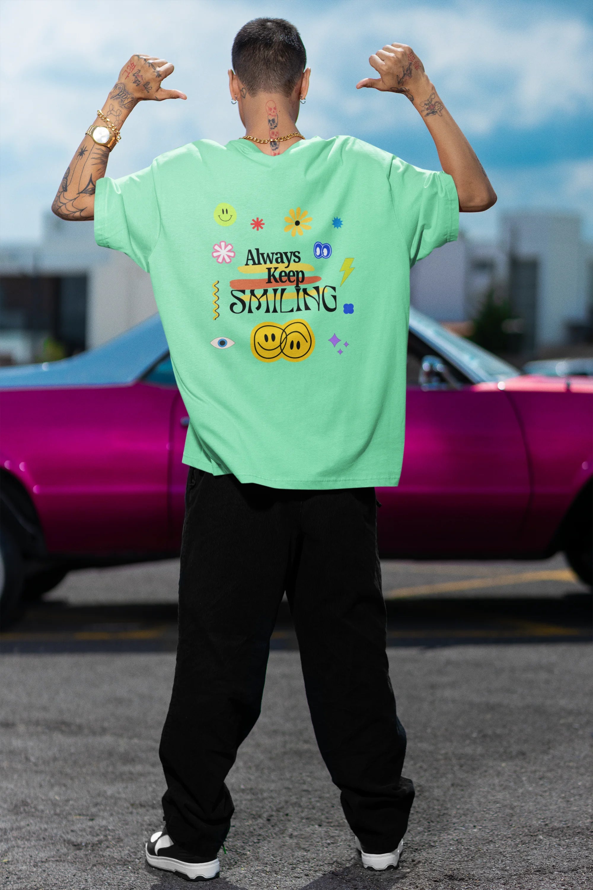 Back view of a male model wearing an oversized mint green t-shirt with a positive message "Keep Smiling" surrounded by colorful flowers and a smiley face emoji. Ideal for those who love cheerful clothing and spreading positivity.