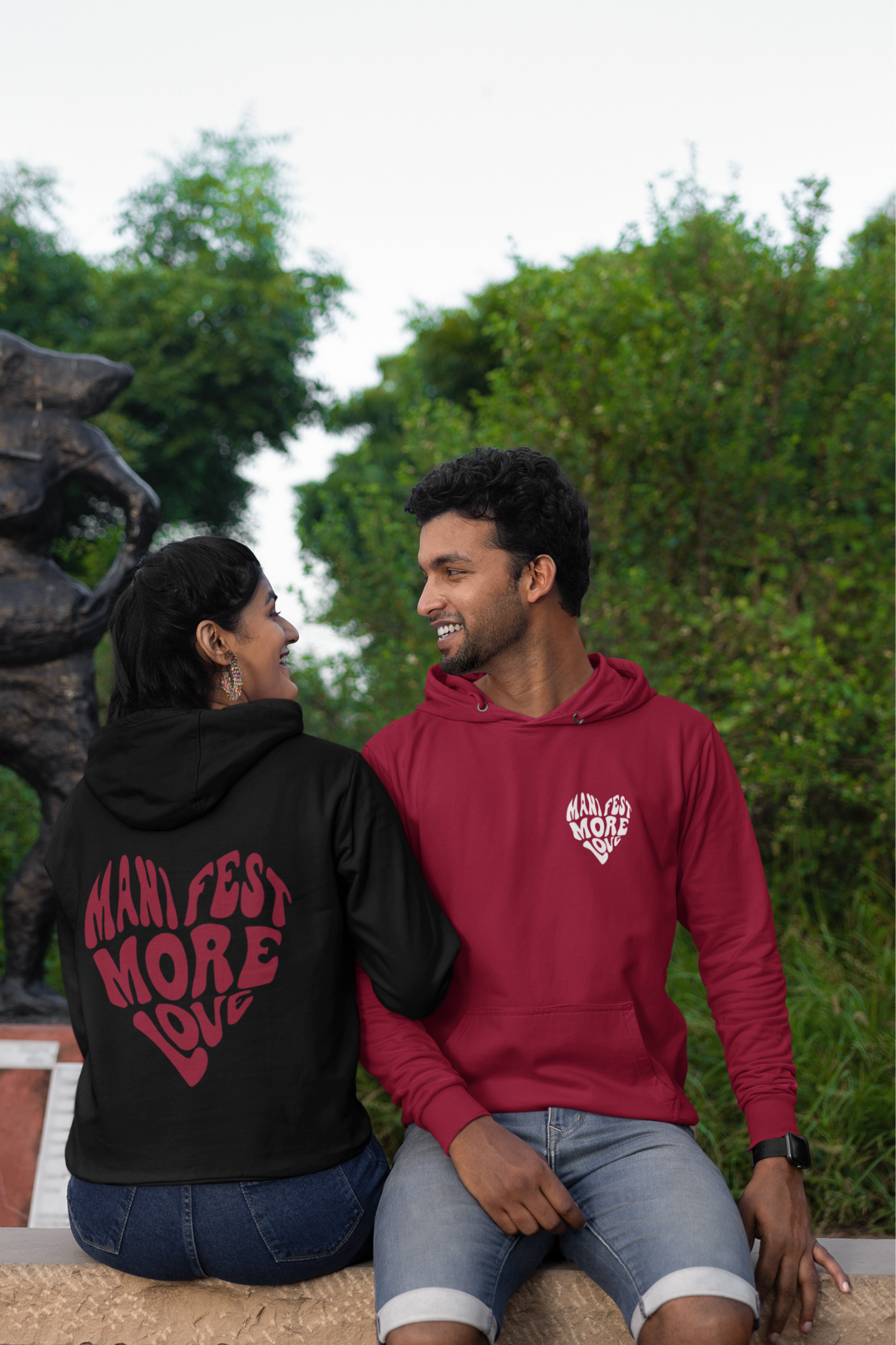 First front and back view of a couple wearing matching hoodies, each with a heart design containing the words "Manifest More Love."