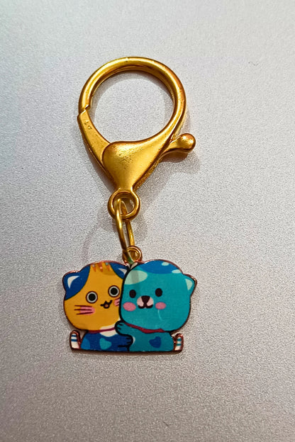 Another view of a couples keychain featuring two separate charms depicting cats hugging each other. The cats' colors may vary, including combinations of pink and blue.