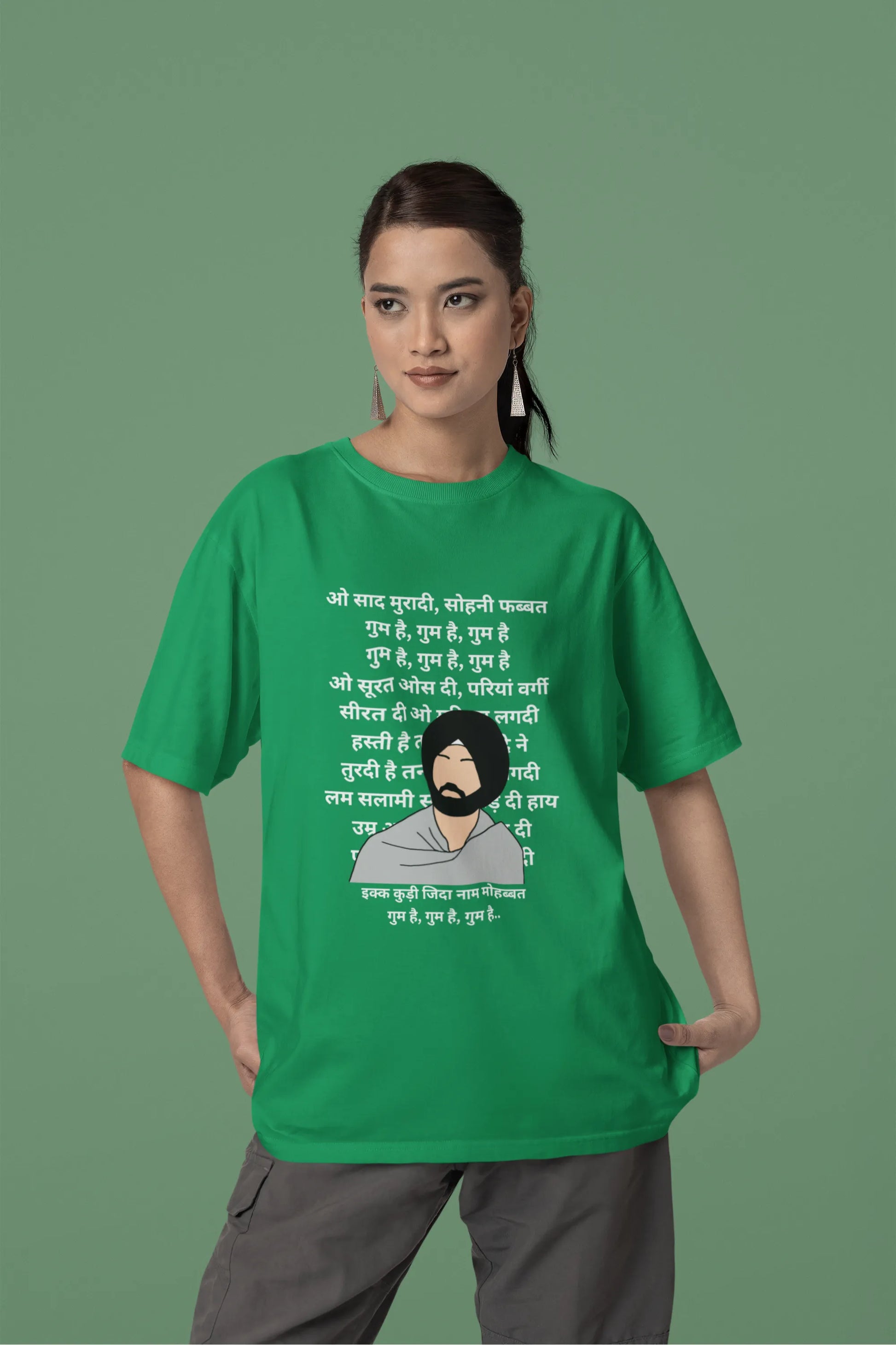 Second front view of a female model wearing a forest green oversized  T-shirt with a Diljit Dosanjh portrait  with lyrics from the song "Ikk Kudi" in the background.