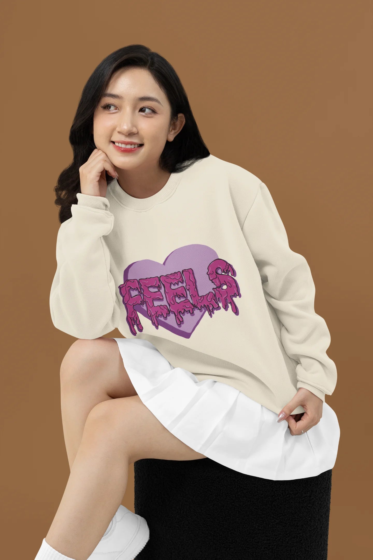 2nd front view of a female model wearing a cream sweatshirt featuring a heart with melting "Feels" text.
