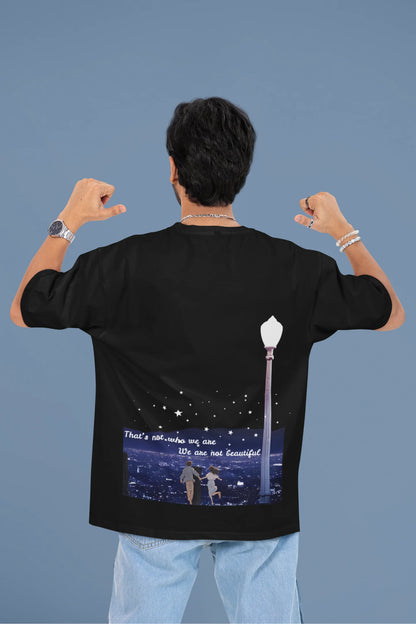 Back view of a male model wearing a black oversized t-shirt featuring a romantic scene of a couple by a lamppost, inspired by Ed Sheeran's "Beautiful People."