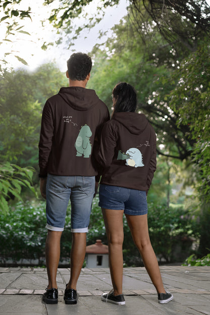 Back view of a couple wearing matching chocolate brown hoodies with a split design. The left side shows one dinosaur pulling the tail of another dinosaur.