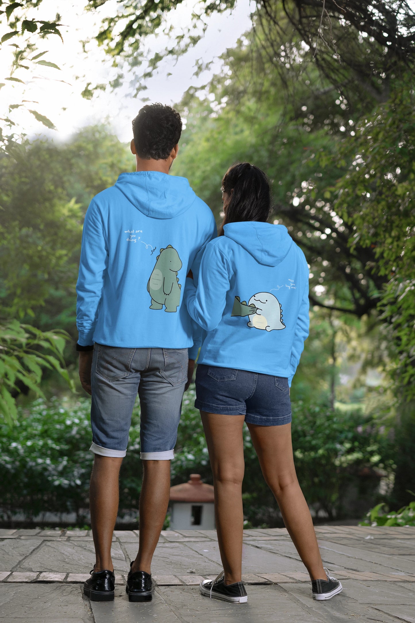 Back view of a couple wearing matching sky blue hoodies with a split design. The left side shows one dinosaur pulling the tail of another dinosaur.