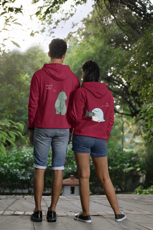 Back view of a couple wearing matching maroon hoodies with a split design. The left side shows one dinosaur pulling the tail of another dinosaur.
