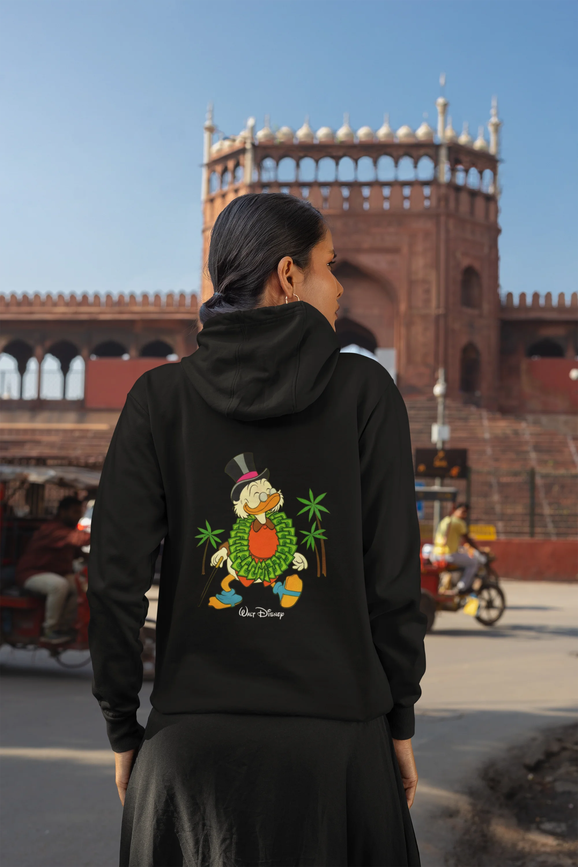 Back view of a female model wearing a black hoodie with a Disney Uncle Scrooge design, showing dollar bills raining down on him.