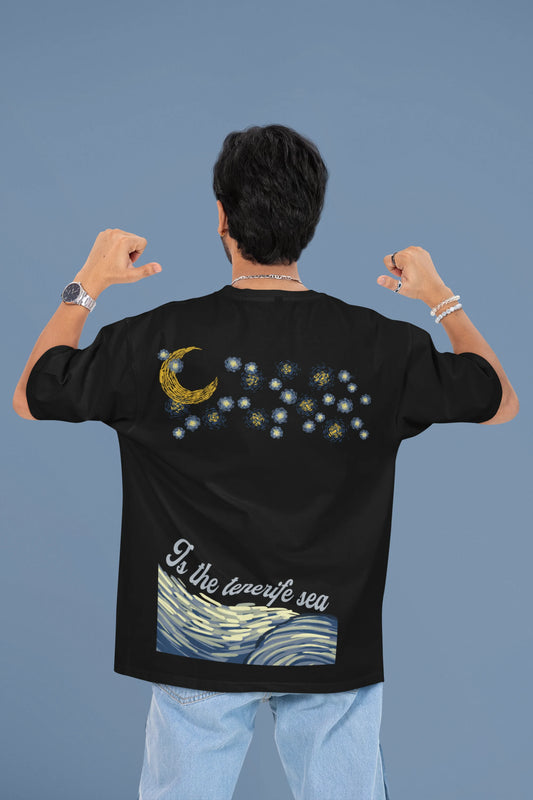 Back view of a male model wearing a black oversized t-shirt featuring a beach at night with a starry sky above, inspired by Ed Sheeran's "Tenerife Sea."