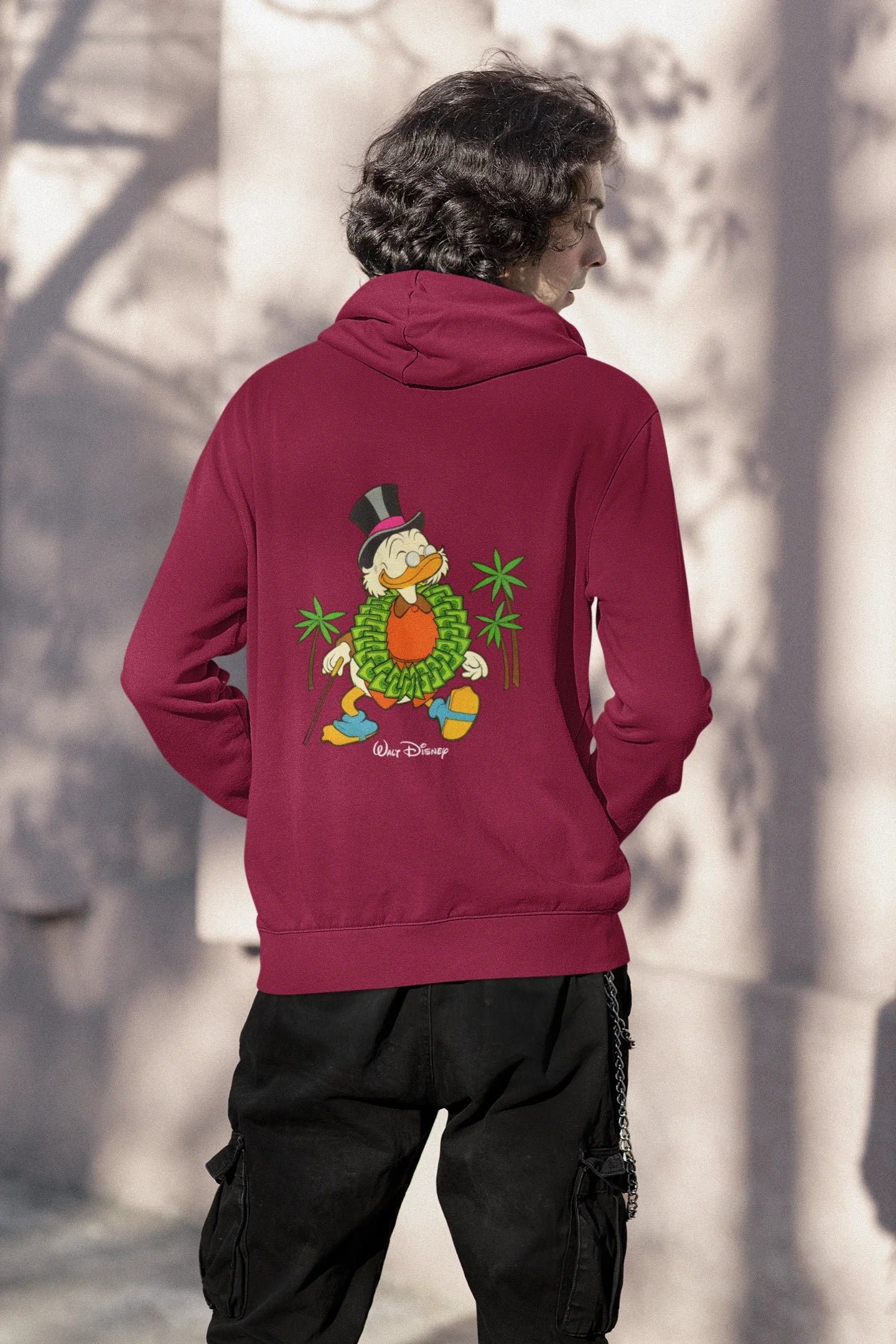 Back view of a male model wearing a maroon hoodie with a Disney Uncle Scrooge design, showing dollar bills raining down on him.