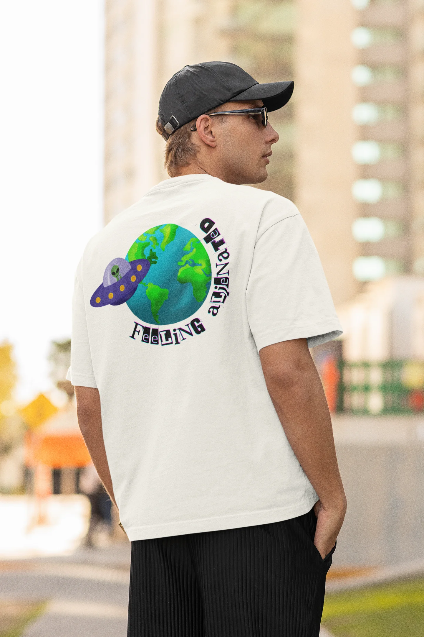 Back view of a male model wearing a off-white oversized t-shirt featuring a fun "Alienated" design with elements of spaceships, UFOs, and a hint of extraterrestrial life.