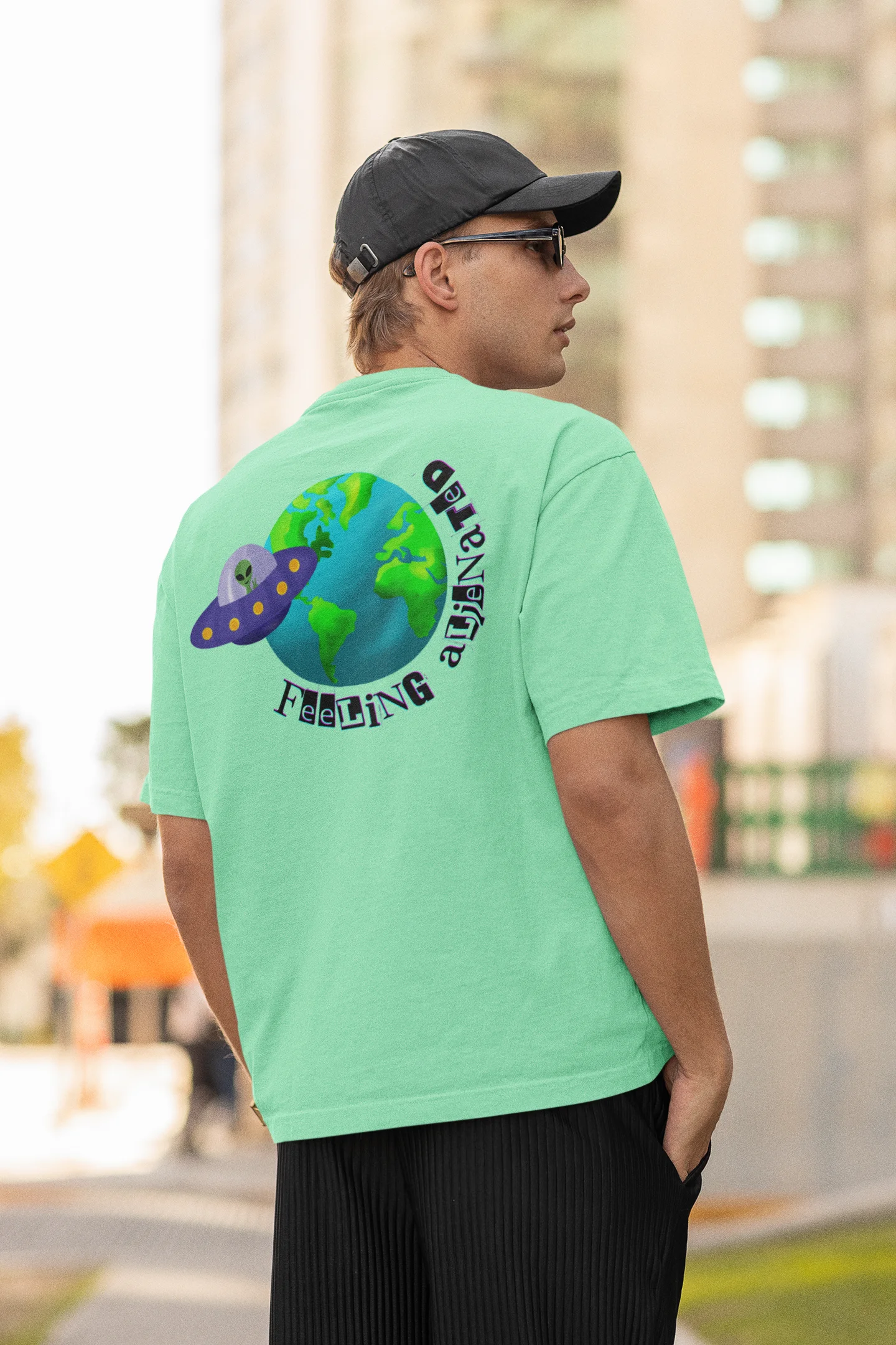 Back view of a male model wearing a mint green oversized t-shirt featuring a fun "Alienated" design with elements of spaceships, UFOs, and a hint of extraterrestrial life.