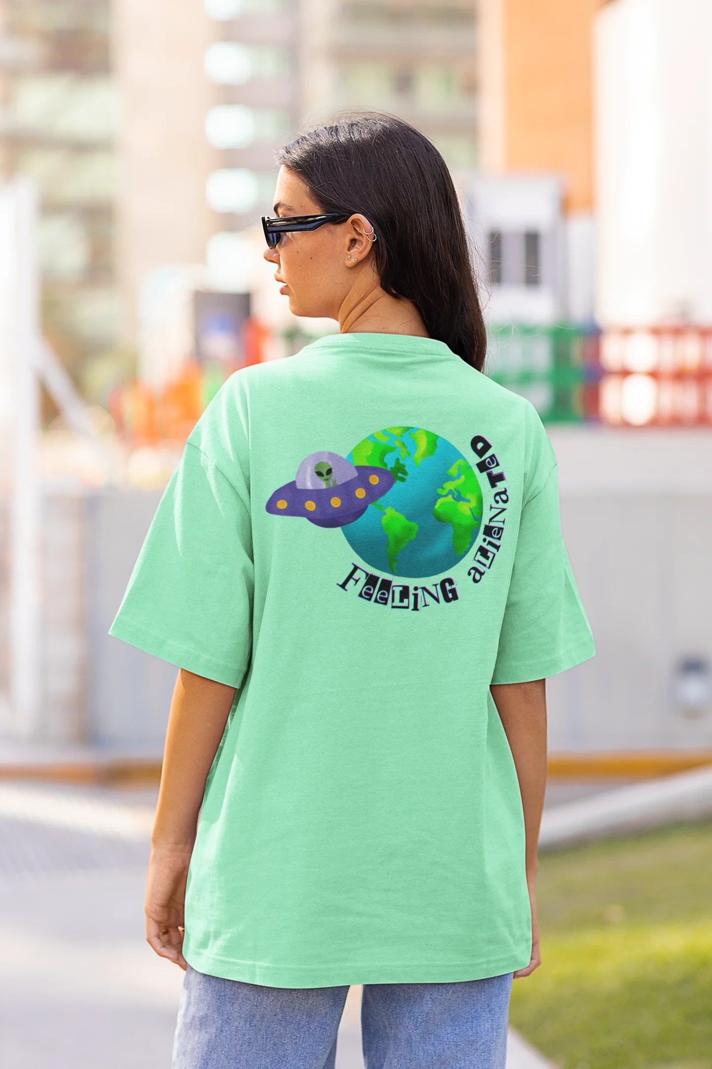 Back view of a female model wearing a mint green oversized t-shirt featuring a fun "Alienated" design with elements of spaceships, UFOs, and a hint of extraterrestrial life.