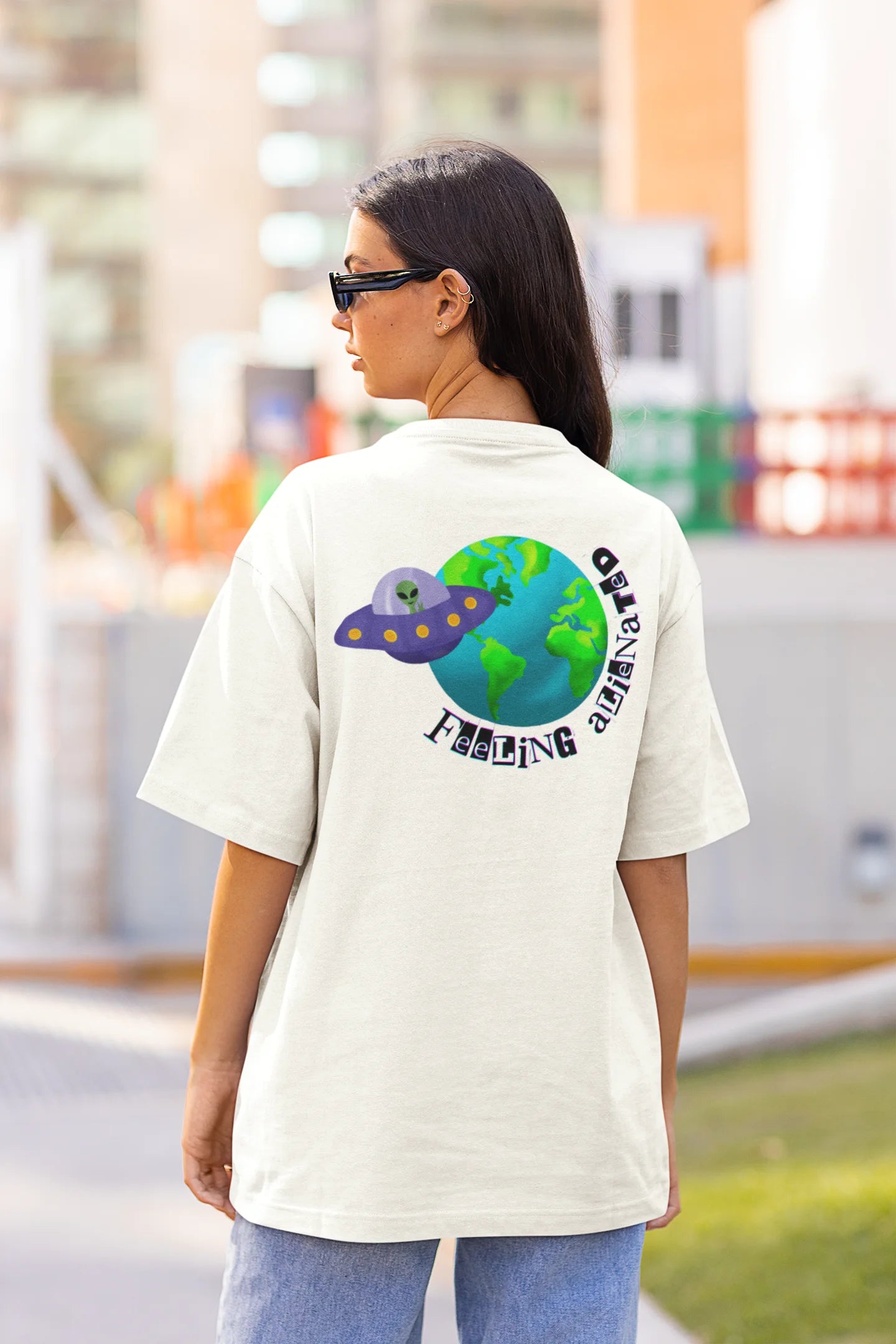 Back view of a female model wearing a off-white oversized t-shirt featuring a fun "Alienated" design with elements of spaceships, UFOs, and a hint of extraterrestrial life.