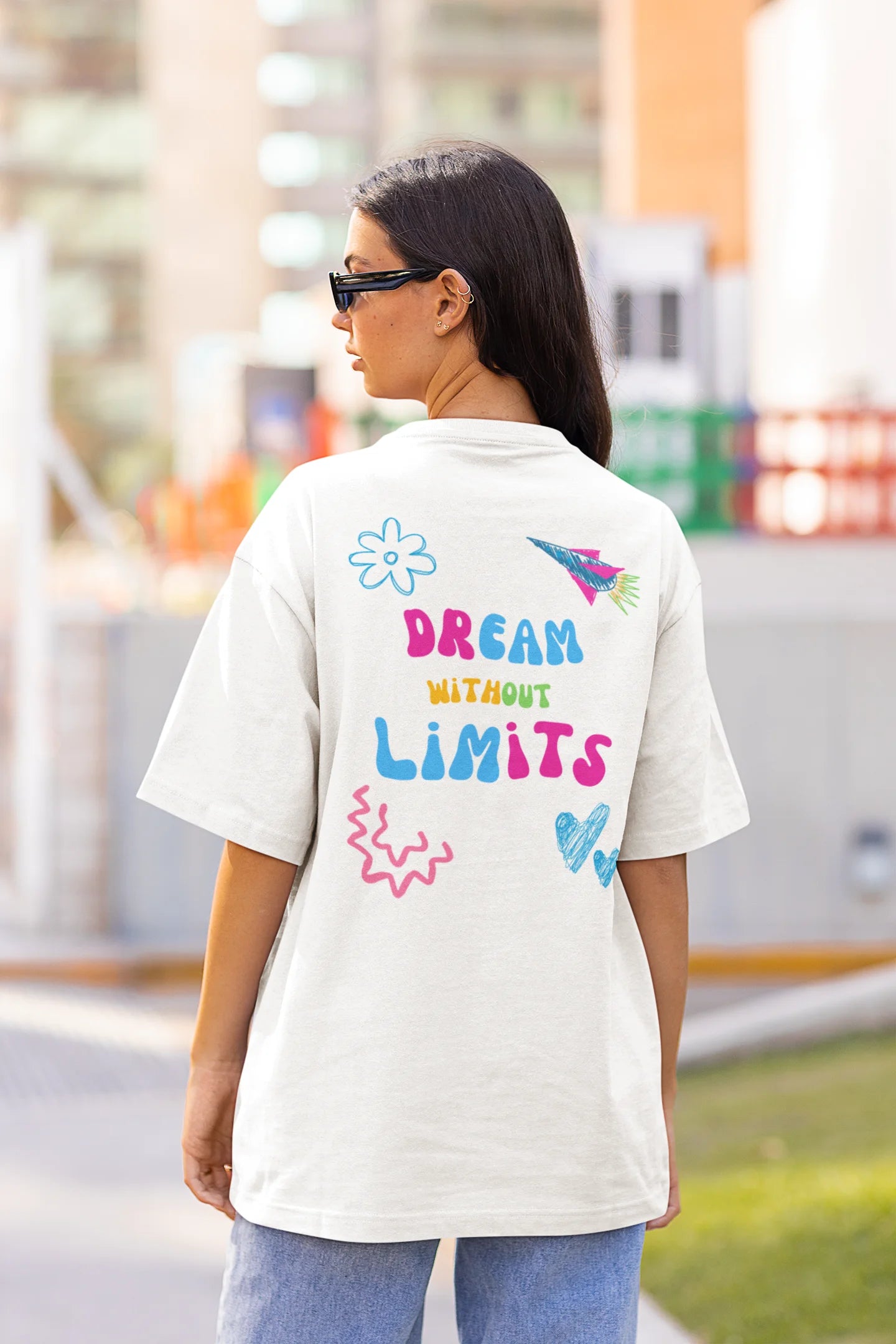 Back view of a female model wearing an off-white oversized t-shirt featuring colorful "Dream Without Limits" text, offering an inspiring and uplifting aesthetic.