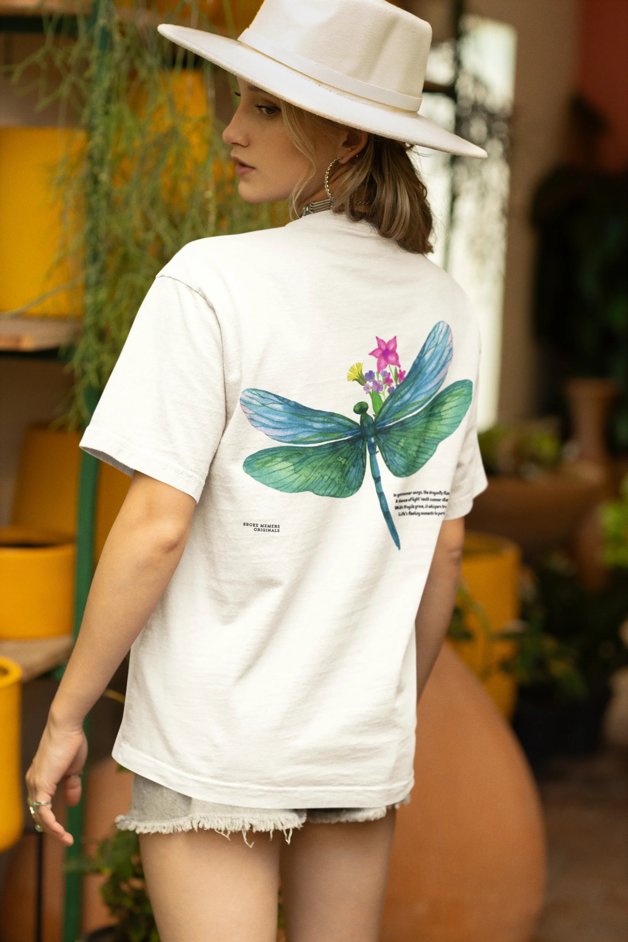 Back view of a female model wearing an off-white oversized t-shirt featuring a nature-inspired design with a blue dragonfly perched on flowers amidst green grass. Ideal for those who love springtime aesthetics and dragonfly imagery.