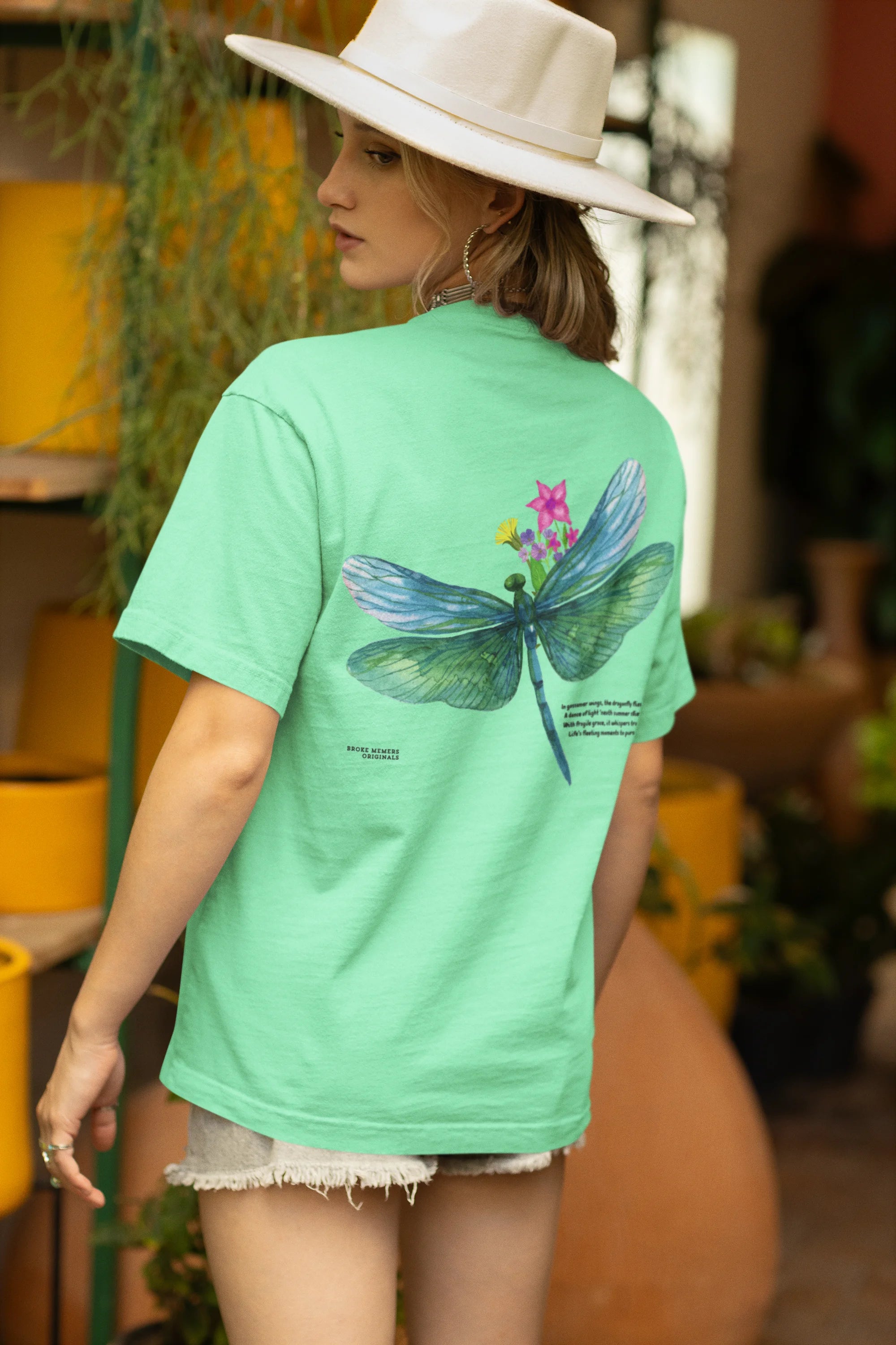 Back view of a female model wearing a mint green oversized t-shirt featuring a nature-inspired design with a blue dragonfly perched on flowers amidst green grass. Ideal for those who love springtime aesthetics and dragonfly imagery.