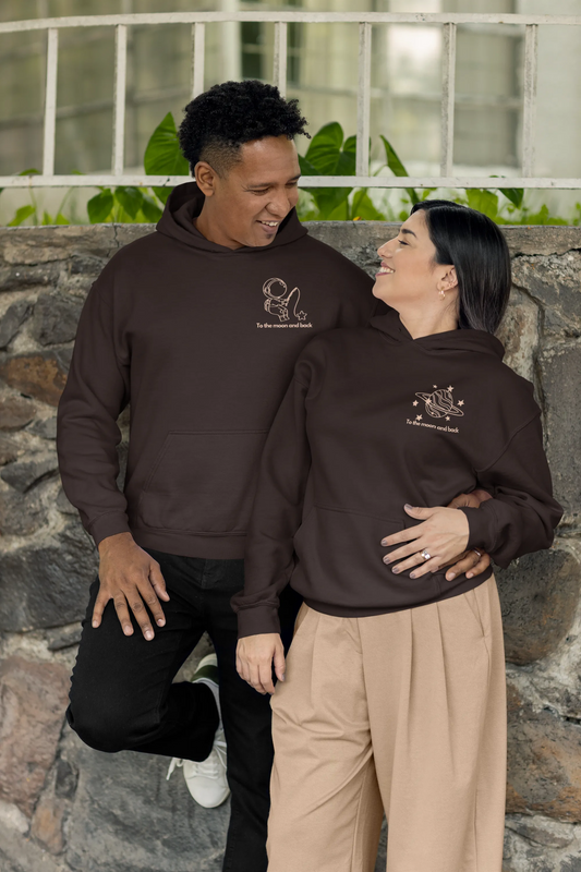 2nd front view of a couple wearing matching chocolate brown hoodies, one with an astronaut design and the other with a planet design, along with the text "To the Moon and Back."