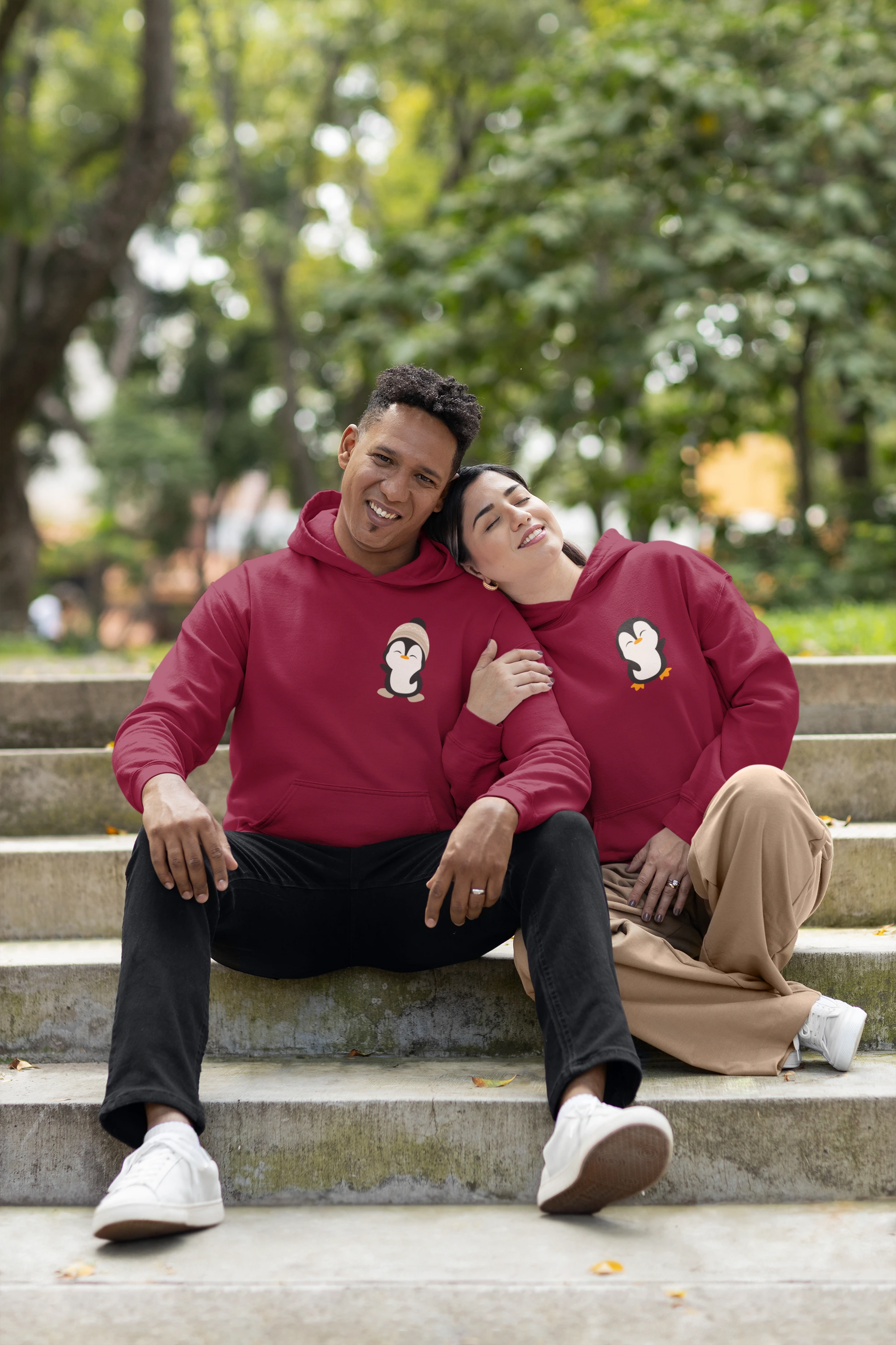 First front view of a couple wearing matching maroon hoodies with a design of two penguins wearing woolen hats.