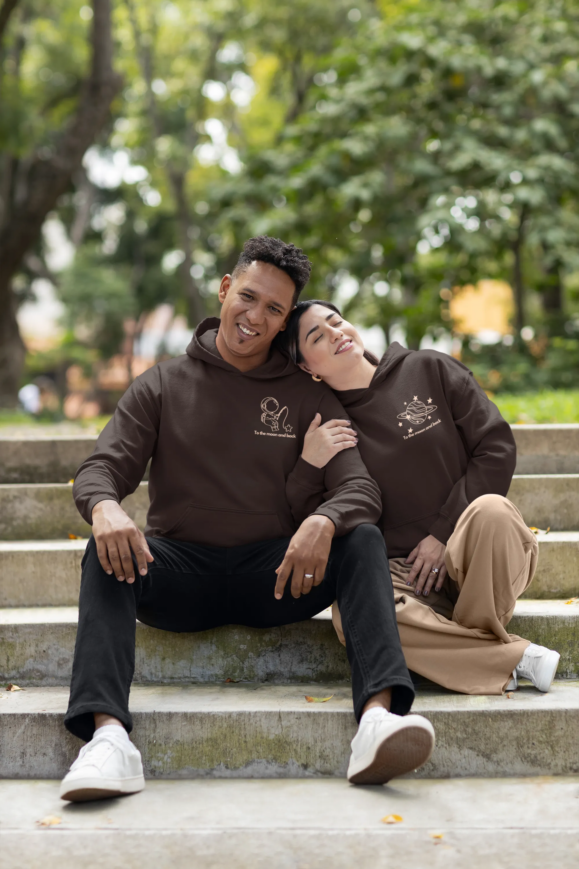 3rd front view of a couple wearing matching chocolate brown hoodies, one with an astronaut design and the other with a planet design, along with the text "To the Moon and Back."