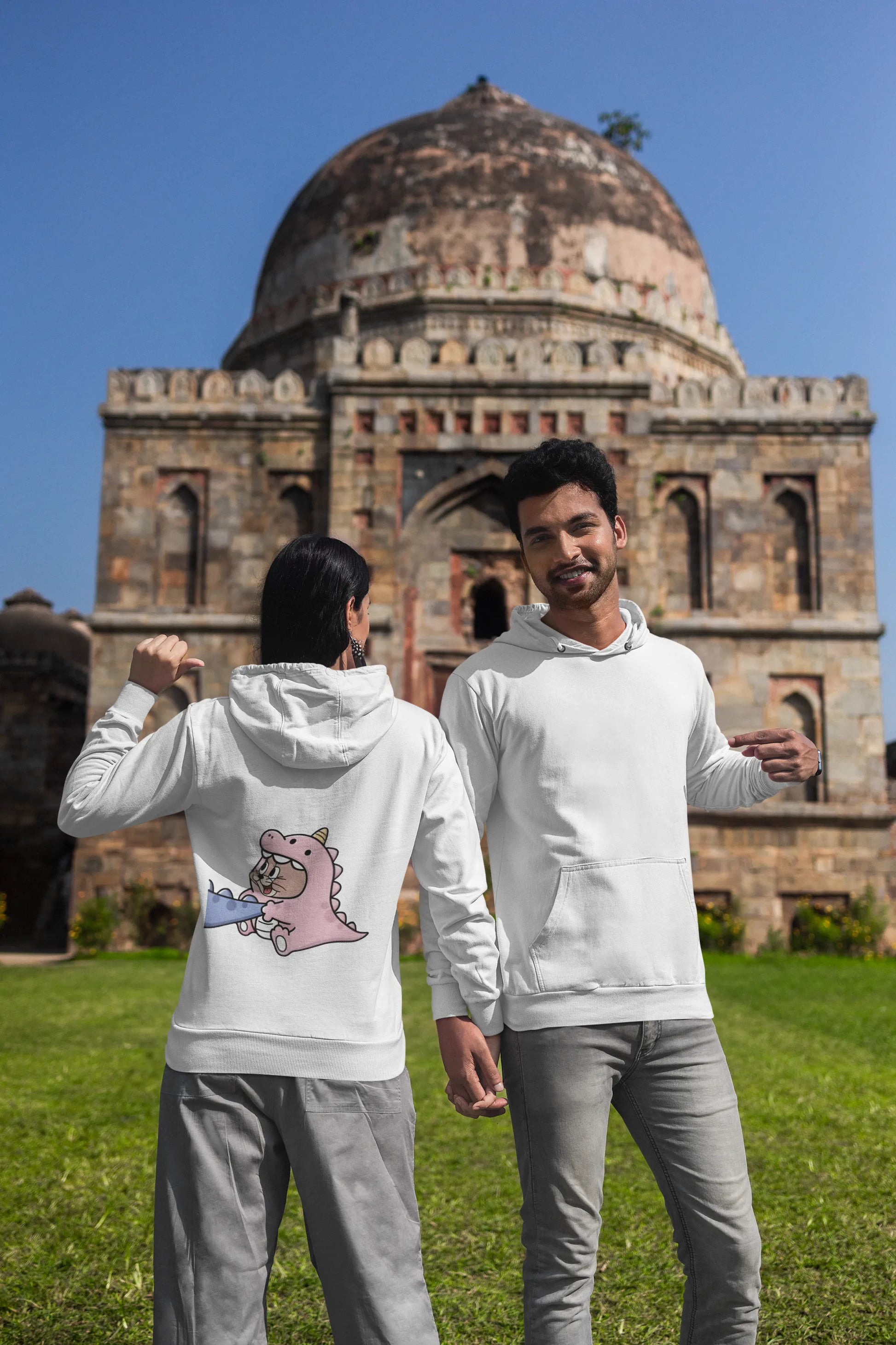 Second back and front view of a couple wearing matching cream hoodies, one with Tom the cat and the other with Jerry the mouse, both dressed in dinosaur costumes.