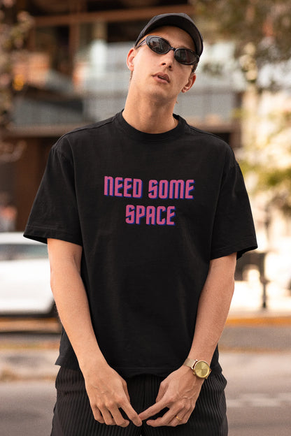 First front view image of a male model wearing a black oversized t-shirt with a bold graphic that reads "Need Some Space" on the front. The design incorporates subtle hints of the Milky Way galaxy and the solar system on the back.