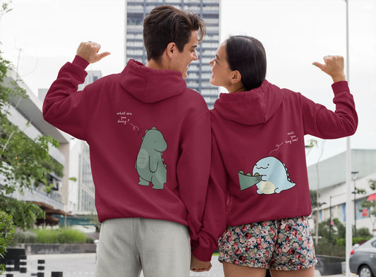 Close up back view of a couple wearing matching maroon hoodies with a split design. The left side shows one dinosaur pulling the tail of another dinosaur.