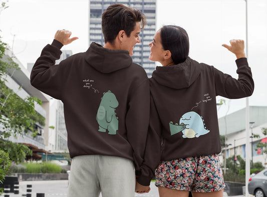 Close up back view of a couple wearing matching chocolate brown hoodies with a split design. The left side shows one dinosaur pulling the tail of another dinosaur.