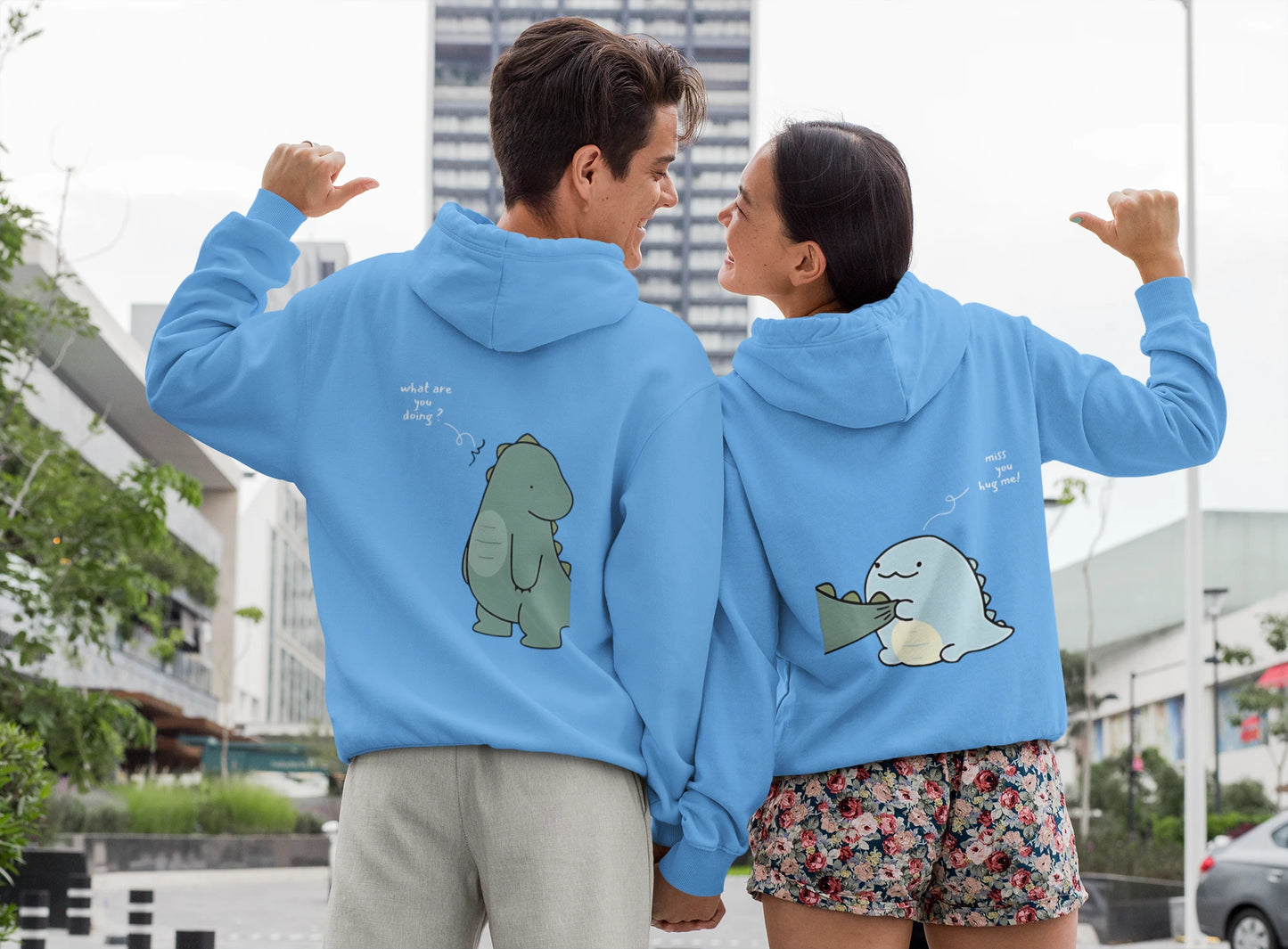 Close up back view of a couple wearing matching sky blue hoodies with a split design. The left side shows one dinosaur pulling the tail of another dinosaur.