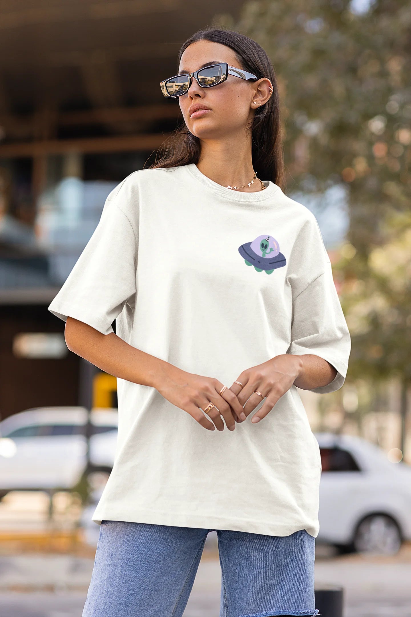 Side view of a female model wearing a off-white oversized t-shirt featuring a fun "Alienated" design with elements of spaceships, UFOs, and a hint of extraterrestrial life.