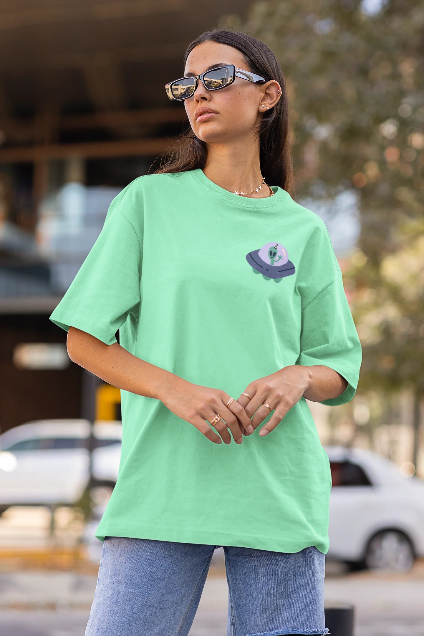 Side view of a female model wearing a mint green oversized t-shirt featuring a fun "Alienated" design with elements of spaceships, UFOs, and a hint of extraterrestrial life.