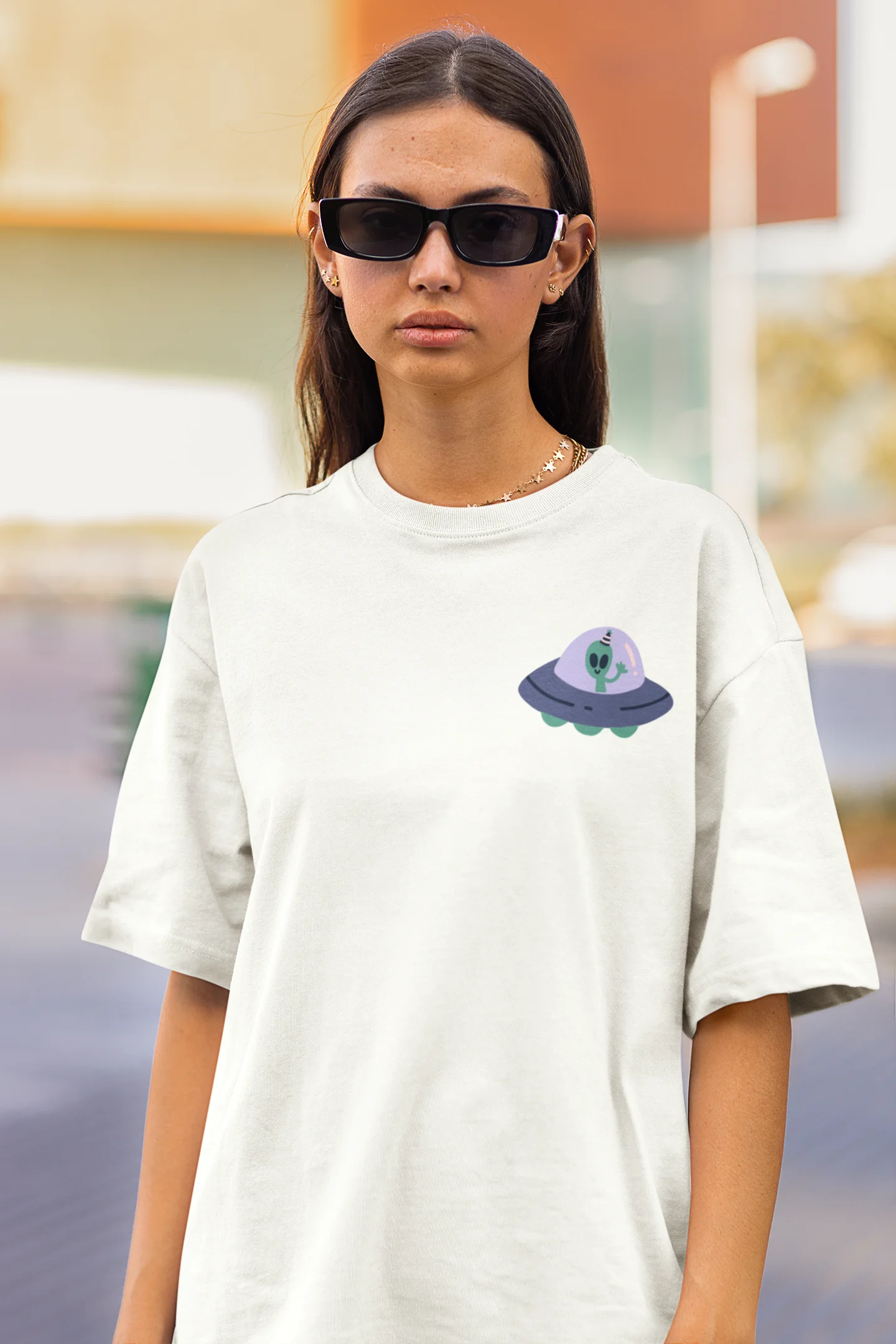 Close up front view of a female model wearing a off-white oversized t-shirt featuring a fun "Alienated" design with elements of spaceships, UFOs, and a hint of extraterrestrial life.