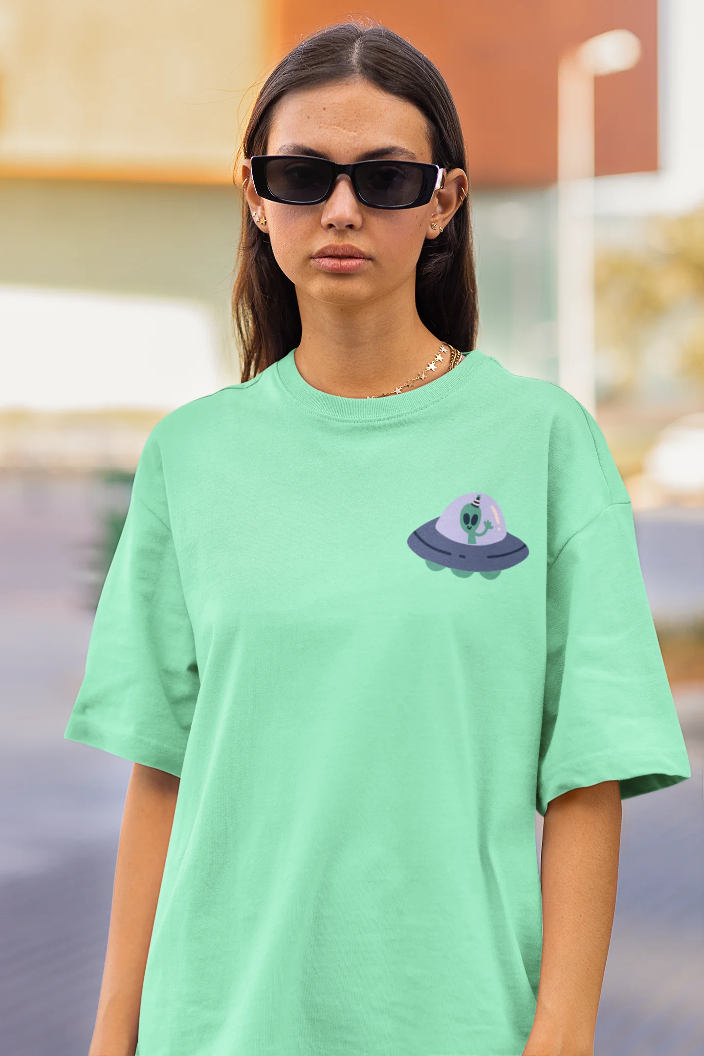 Close up front view of a female model wearing a mint green oversized t-shirt featuring a fun "Alienated" design with elements of spaceships, UFOs, and a hint of extraterrestrial life.